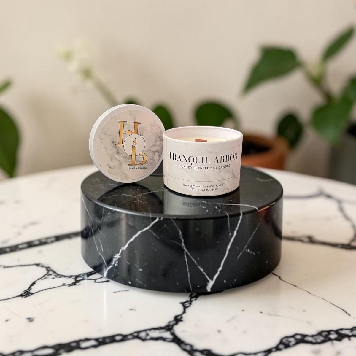 A scented soy candle labeled 'TRANQUIL ARBOR' sits on a round black marble pedestal next to its matching marble lid, which features a gold monogram logo. The setup is on a white marble table with a blurred indoor background containing plants.